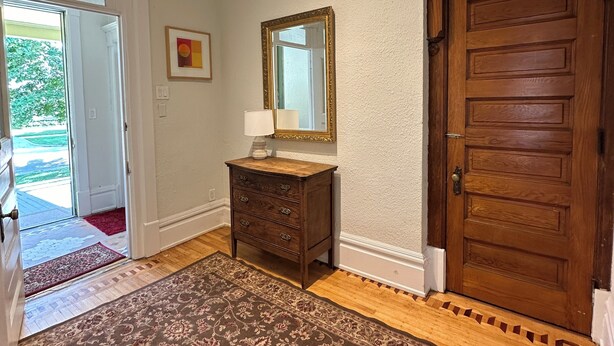 A hallway with a wooden floor, a patterned rug, a wooden dresser with a mirror, and a wooden door.