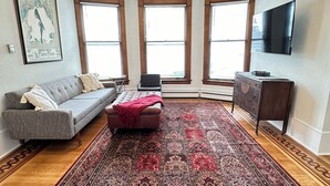 A living room with a chandelier, a gray sofa, a red ottoman, a patterned rug, and a wooden floor.