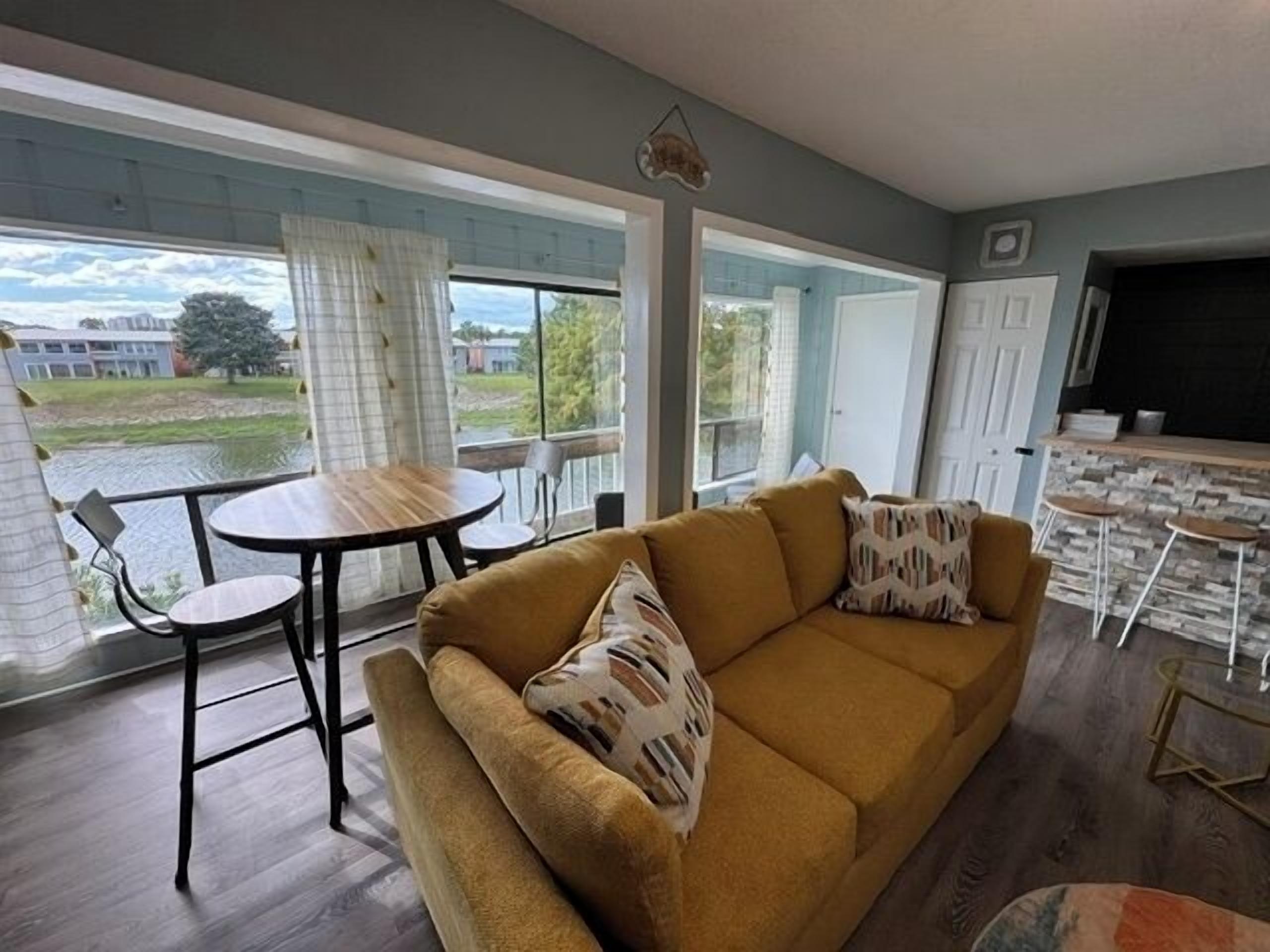 A living room with a sofa, a small table, and chairs. There is a large window with curtains, a door, and a kitchen area with a counter and stools.