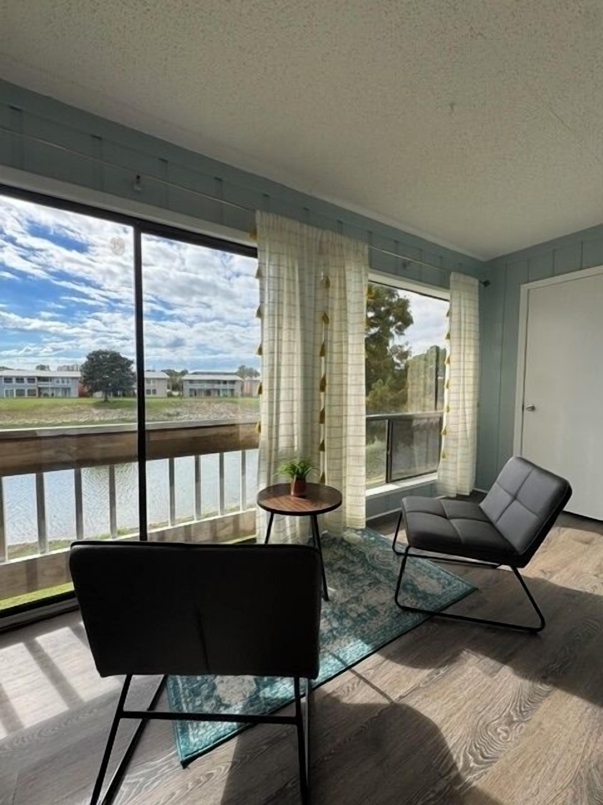 A room with a large window, a black chair, a small table with a plant, and a view of a building and trees.