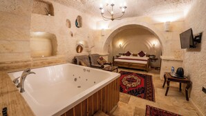 A bathroom with a large bathtub, a sink, and a shower. There is a chair and a small table with a lamp. The walls are made of stone, and there is a decorative rug on the floor. - Library Cave Hotel (Urgup)