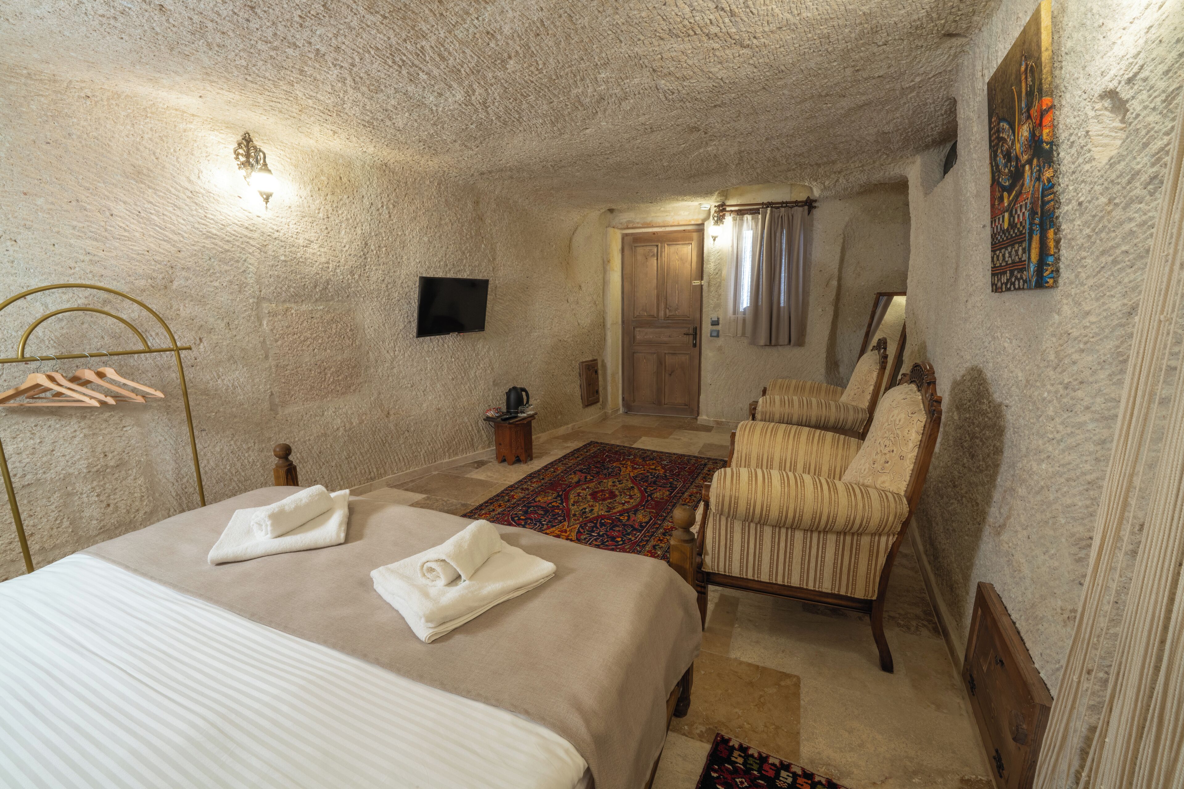 A hotel room with a large bed, two armchairs, a small table, a TV, and a door leading to another room.