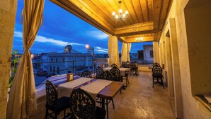 An outdoor dining area with wooden tables and chairs, a view of the cityscape, and a covered ceiling with a chandelier. - Library Cave Hotel (Urgup)