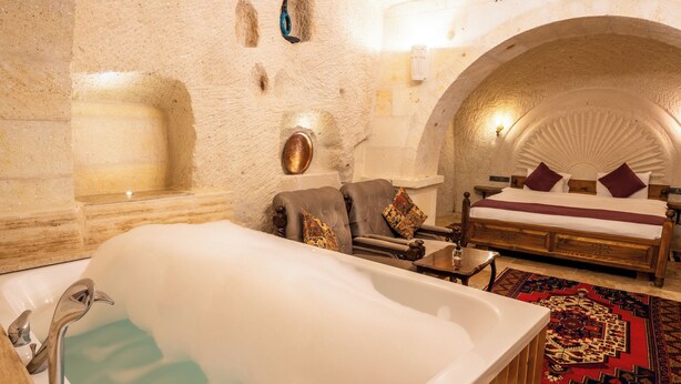 A luxurious room with a large bathtub, a wooden bench, a bed with a red and black patterned rug, and a chandelier. - Library Cave Hotel (Urgup)