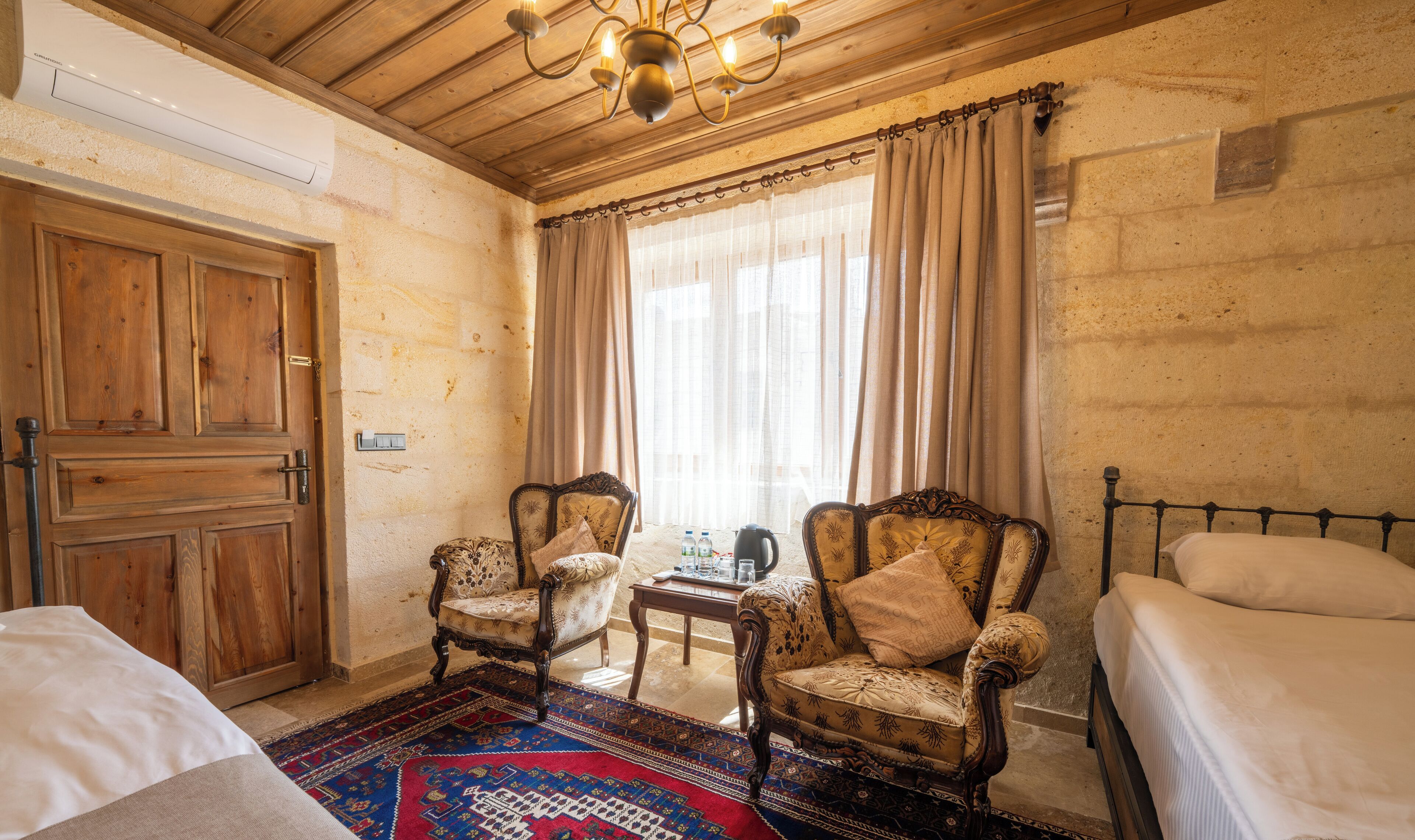 A room with two wooden chairs, a small table, a bed, and a large window with curtains.