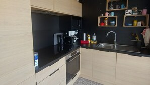 Eine moderne Küche mit hellen Holzschränken, einer schwarzen Arbeitsplatte und einem eingebauten Backofen. Regale mit Küchenutensilien und ein kleiner Spülbereich sind ebenfalls vorhanden.