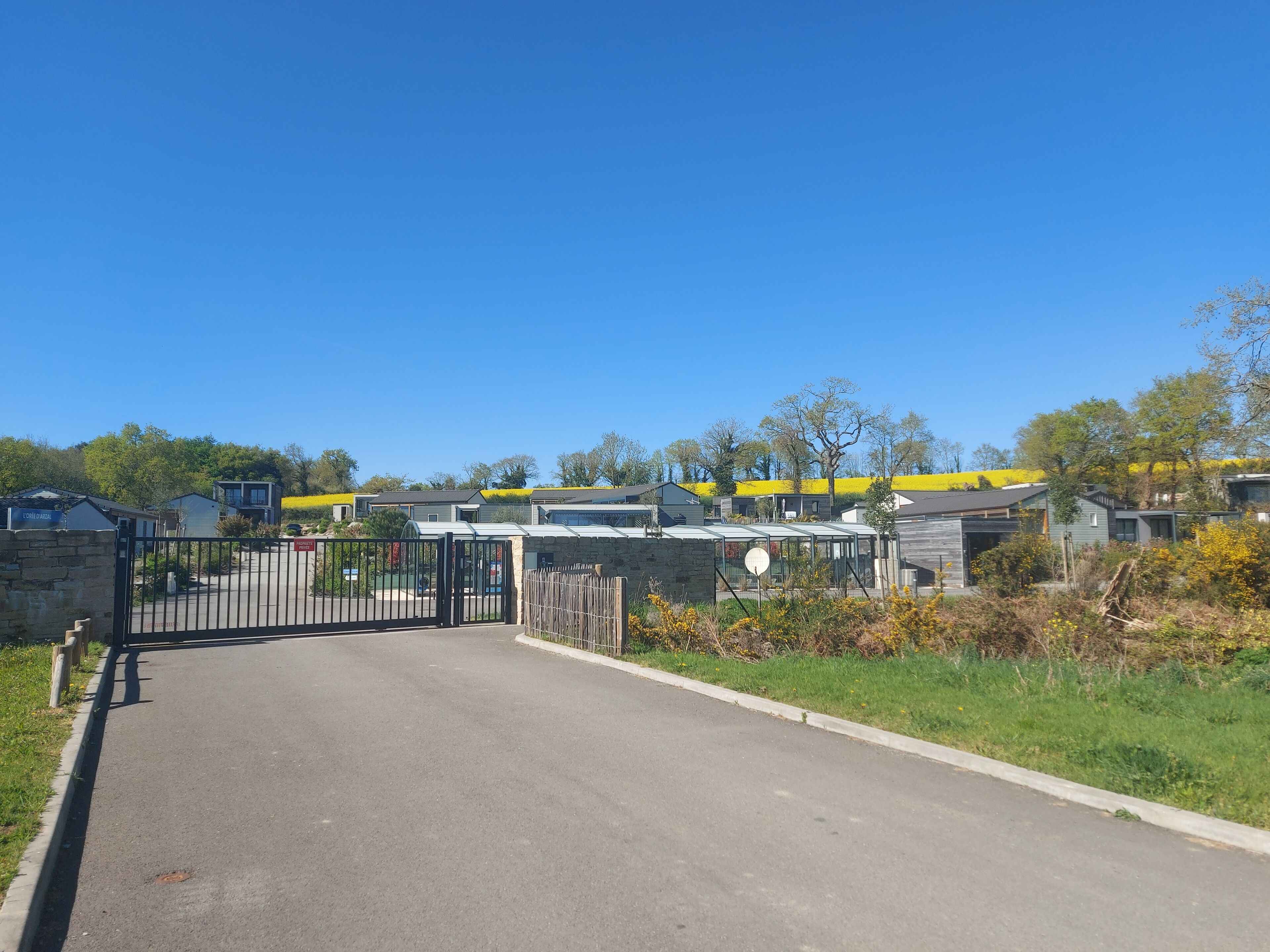 Une entrée fermée par un portail donnant accès à un quartier résidentiel avec des maisons modernes et un ciel d’un bleu éclatant.