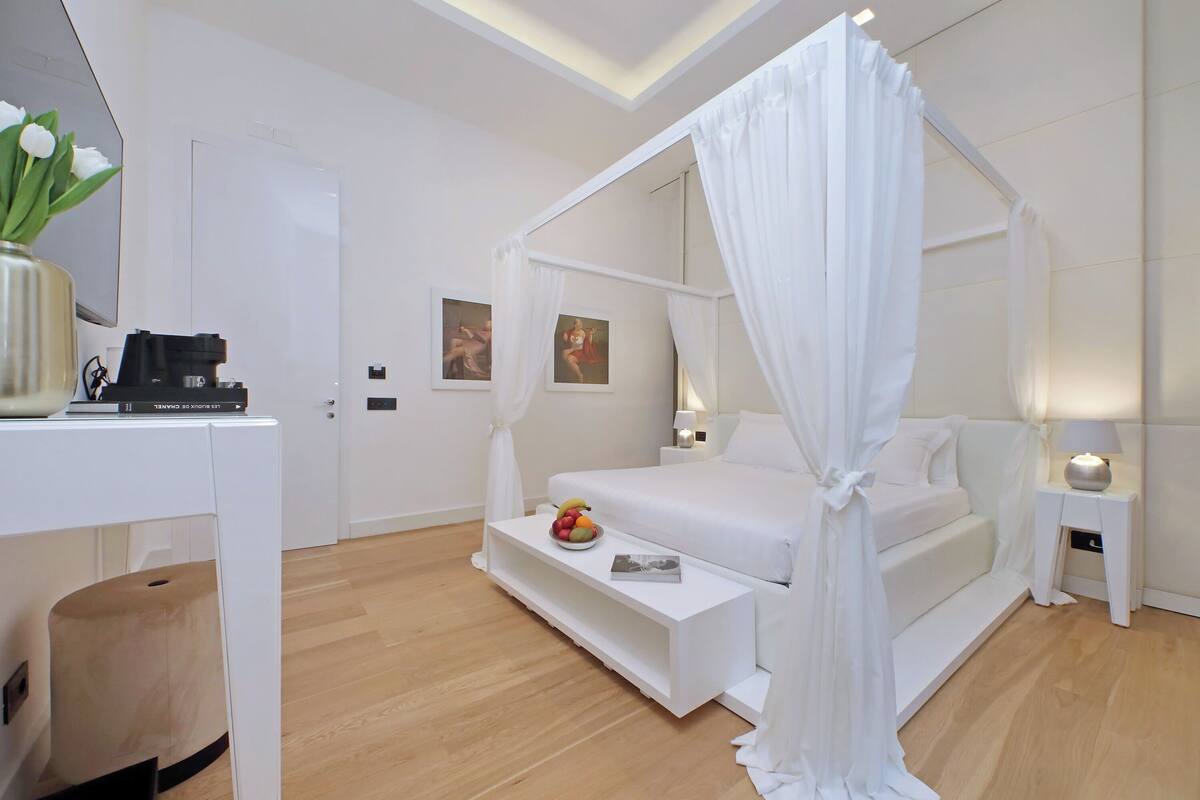 A modern bedroom with a four-poster bed, bedside tables, a nightstand, and a small table with fruit.