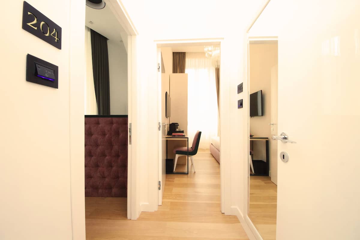 A hotel room with a wooden floor, a tufted brown sofa, a desk with a chair, a television, and a door with a number sign.