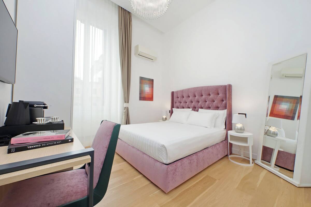 A modern hotel room with a pink upholstered bed, a desk with a laptop, and a mirror.