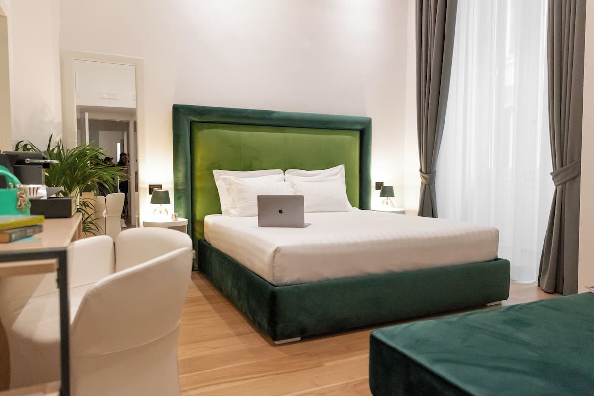A modern hotel room with a green upholstered bed, a laptop on the bed, a desk with a chair, and a bedside table with a lamp.