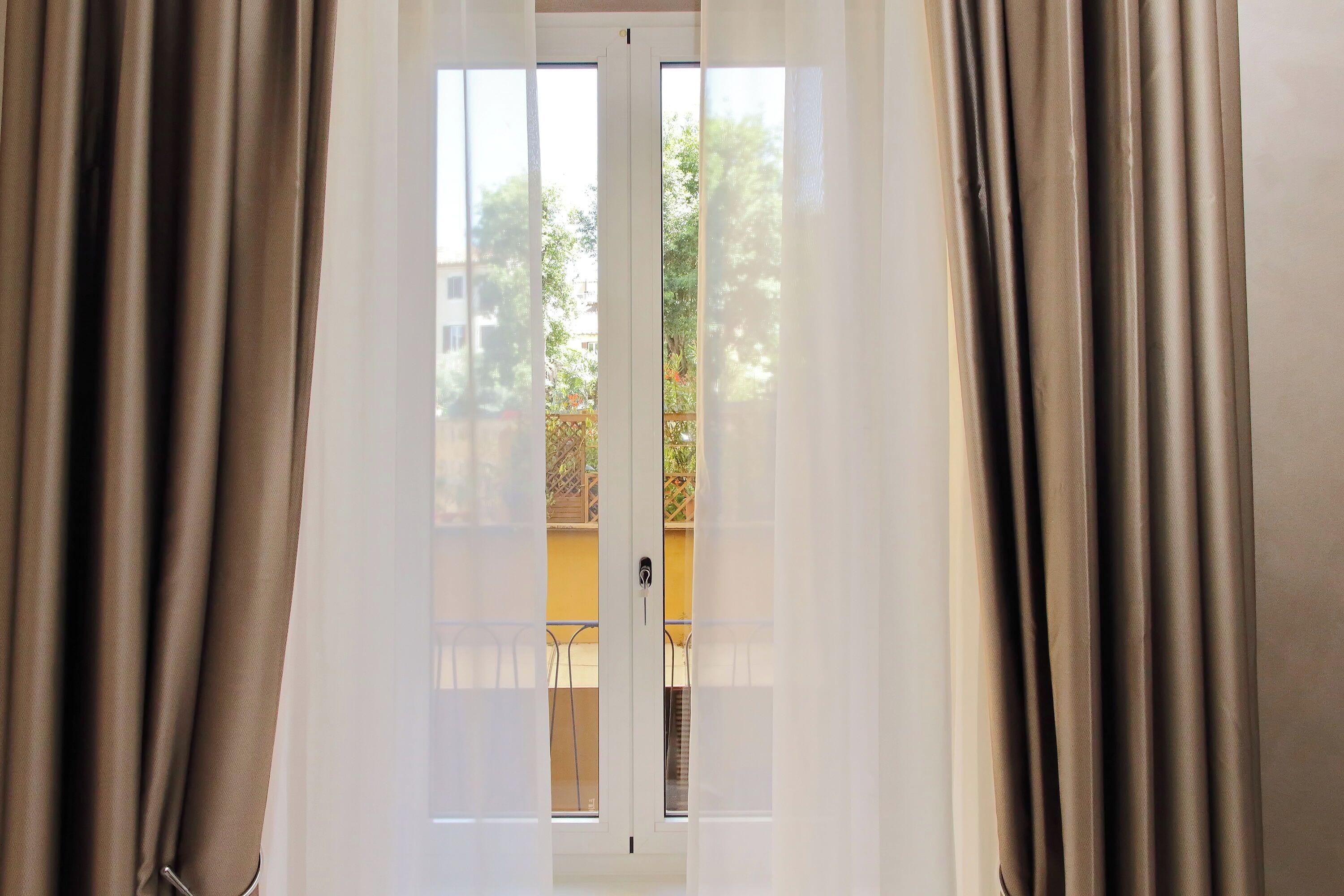 A view through a window with curtains, revealing a balcony with a railing and a yellow wall.