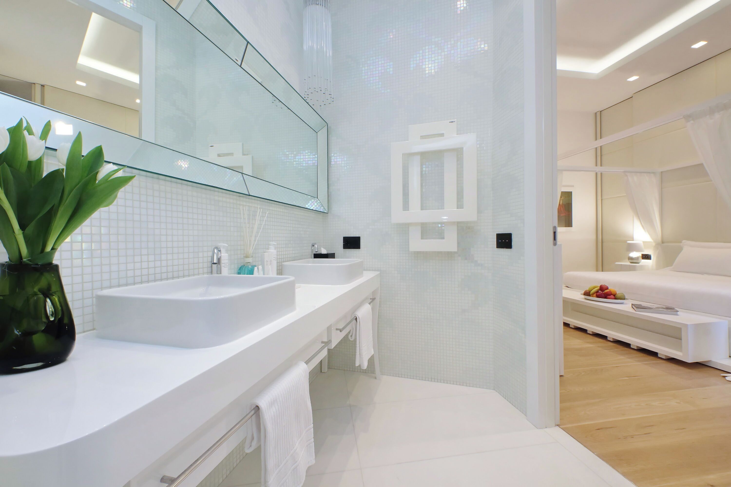 A modern bathroom with two sinks, a large mirror, and a bedside table with a fruit bowl.