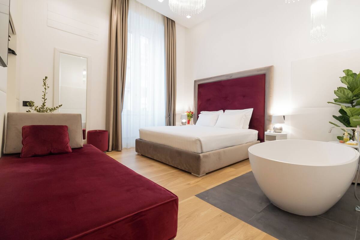 A modern hotel room with a large bed, a round bathtub, and a seating area with a red sofa.