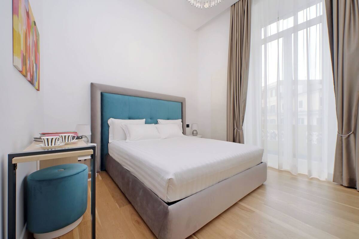 A modern bedroom with a large bed, a bedside table, a lamp, and a view of the city through sheer curtains.