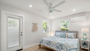 Un dormitorio con cama, ventilador de techo, ventana con persianas y mesita de noche con lámpara.