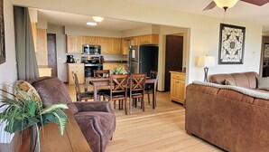 Un salon avec un canapé marron, un coin repas avec des chaises en bois, une cuisine avec des meubles en bois et un ventilateur de plafond.