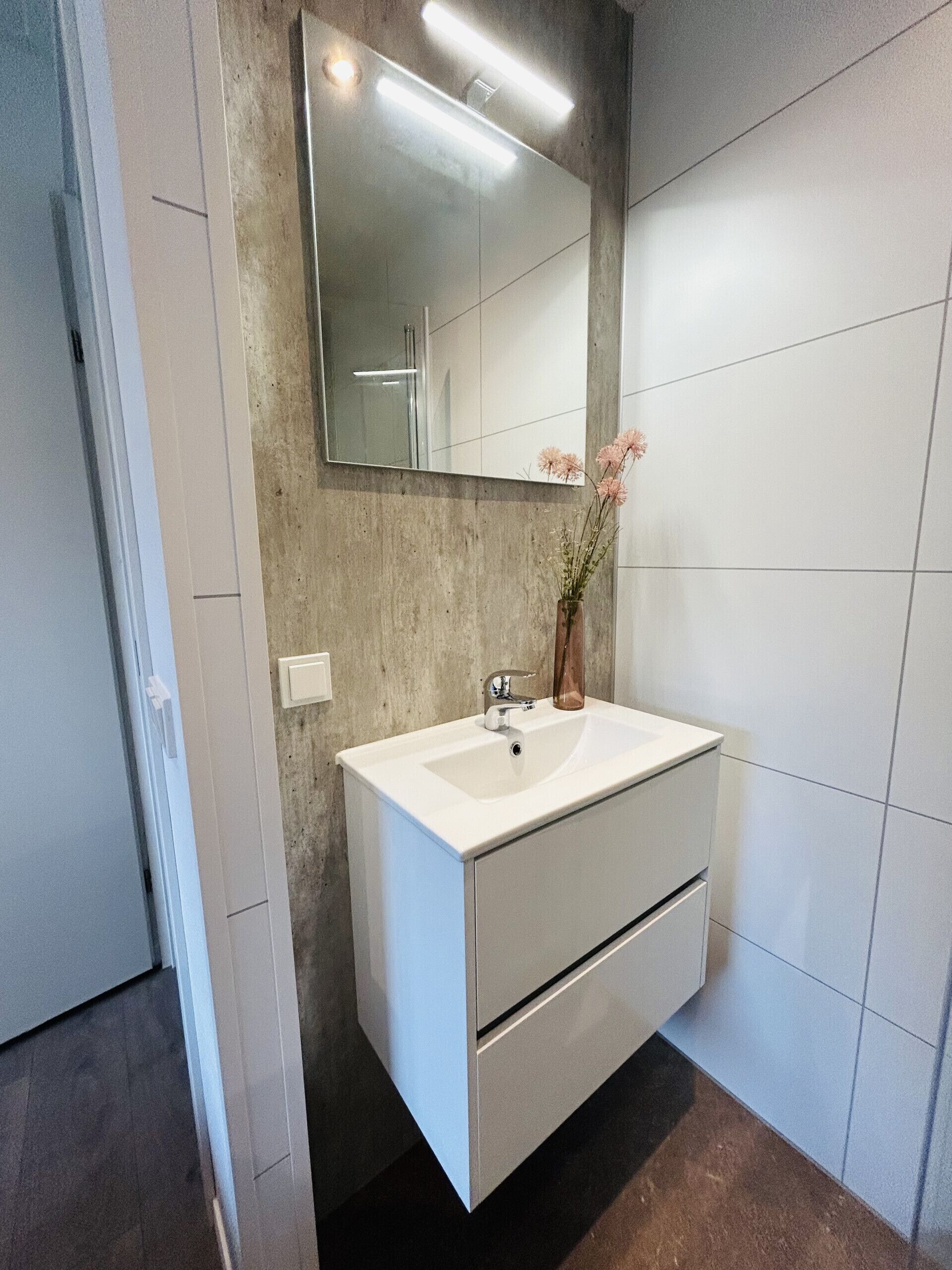 Un bagno moderno con un lavandino bianco, uno specchio e un vaso con fiori.