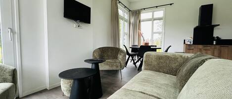 Un salon comprenant un canapé, deux fauteuils, une table basse, un téléviseur à écran plat fixé au mur, et un coin repas avec une table et des chaises.