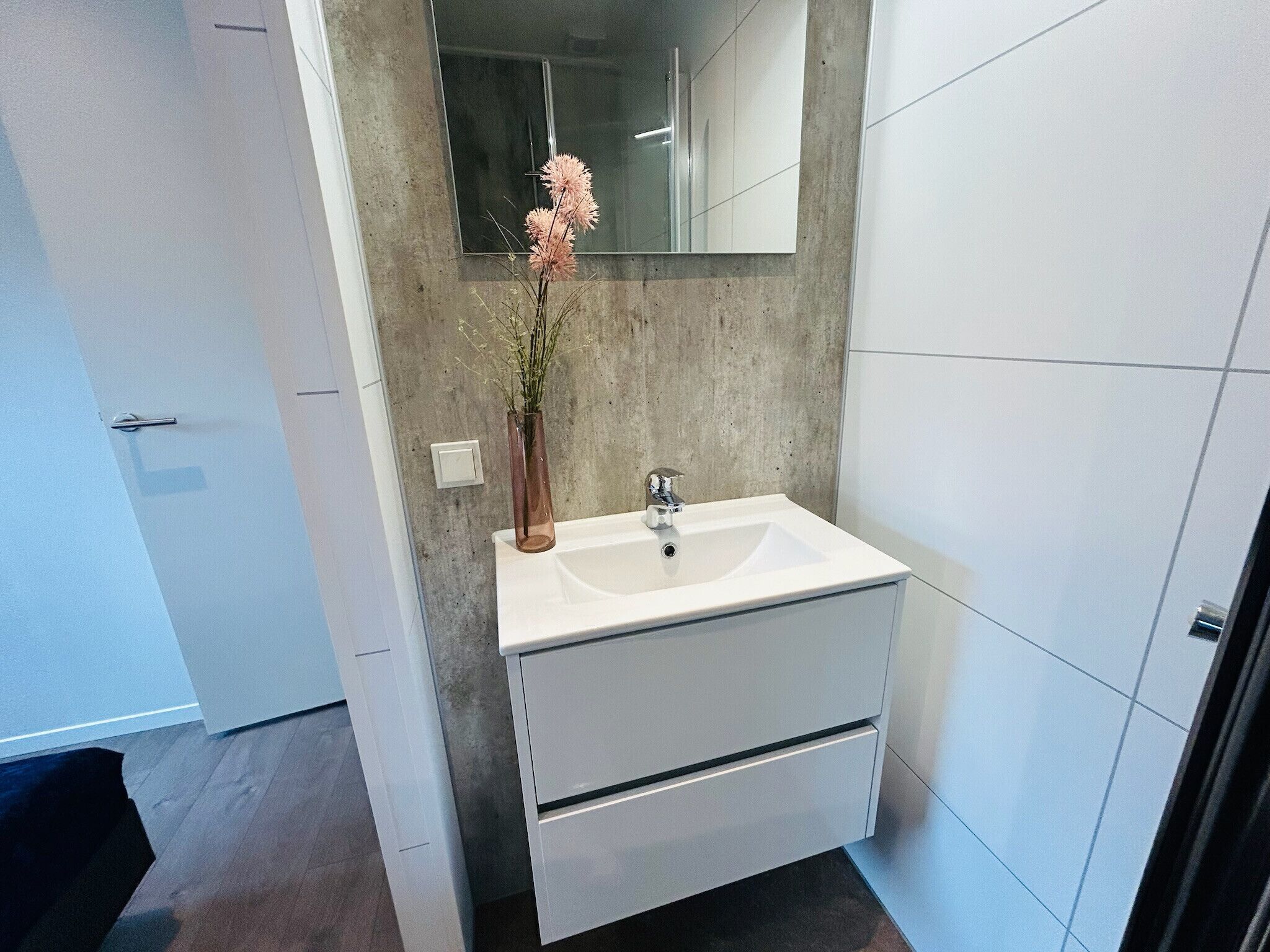 Un bagno moderno con un mobile lavabo bianco, uno specchio grande e un vaso con fiori rosa.