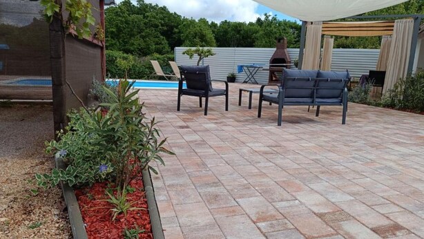 Une terrasse extérieure avec une piscine, deux chaises noires, une pergola en bois et un parterre fleuri.