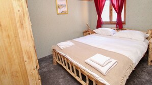 Un lit en bois avec du linge de lit blanc et un plaid beige, une table de chevet avec une lampe, et une fenêtre avec des rideaux rouges.