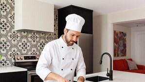 A chef in a white uniform slicing vegetables on a kitchen island. - Artisan's Cove at Water's Edge by Liberty with Golf Simulator (Municipality Of Chatham-Kent)