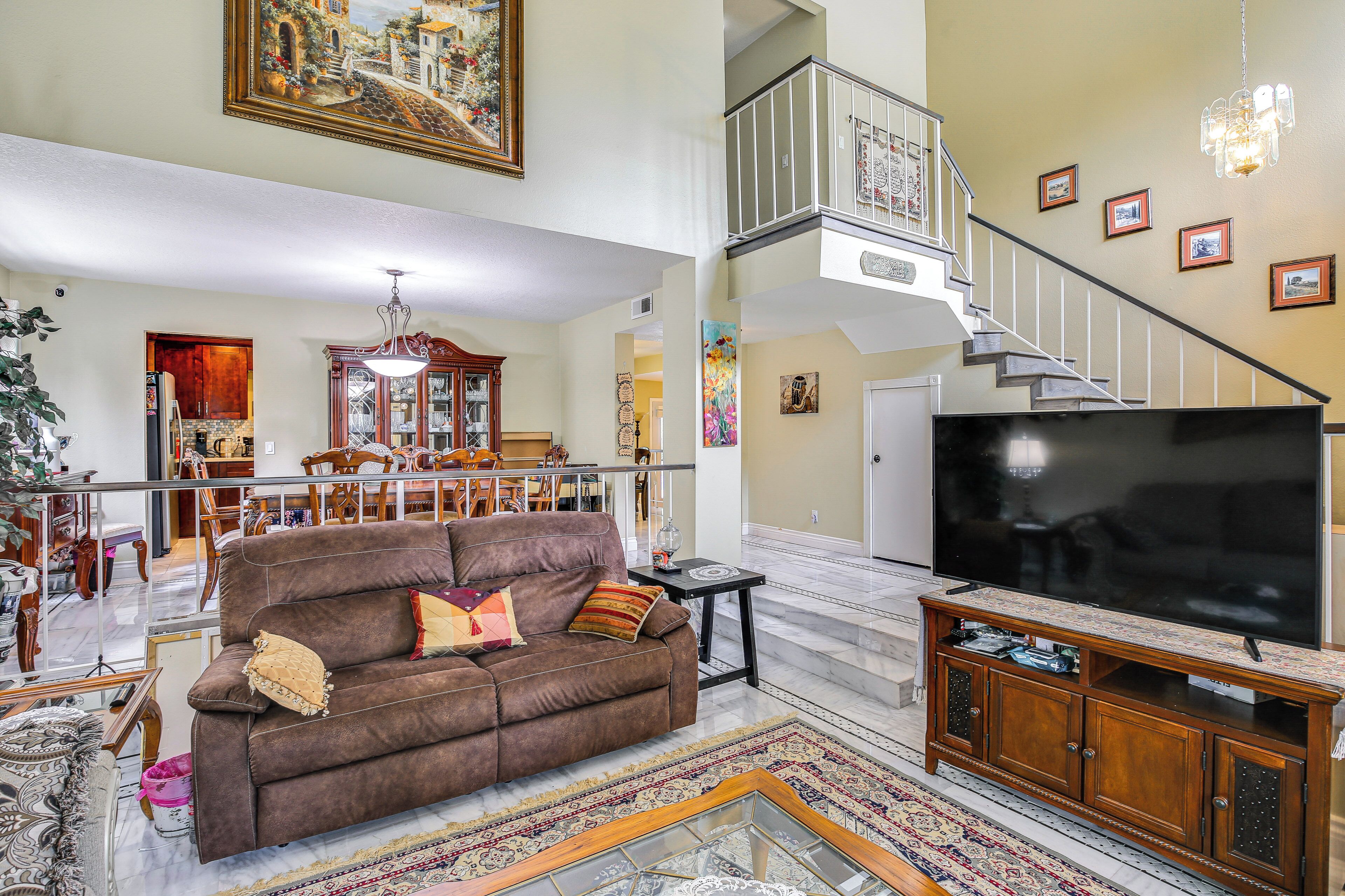 A spacious living room with a brown sofa, a large flat-screen TV, a wooden entertainment unit, and a staircase leading to an upper level.