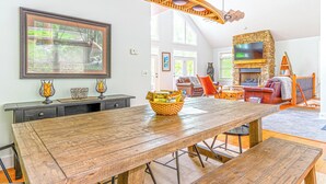 Ein geräumiges Wohnzimmer mit einem großen Esstisch aus Holz, einem Kamin aus Stein und einem an der Wand befestigten Fernseher.