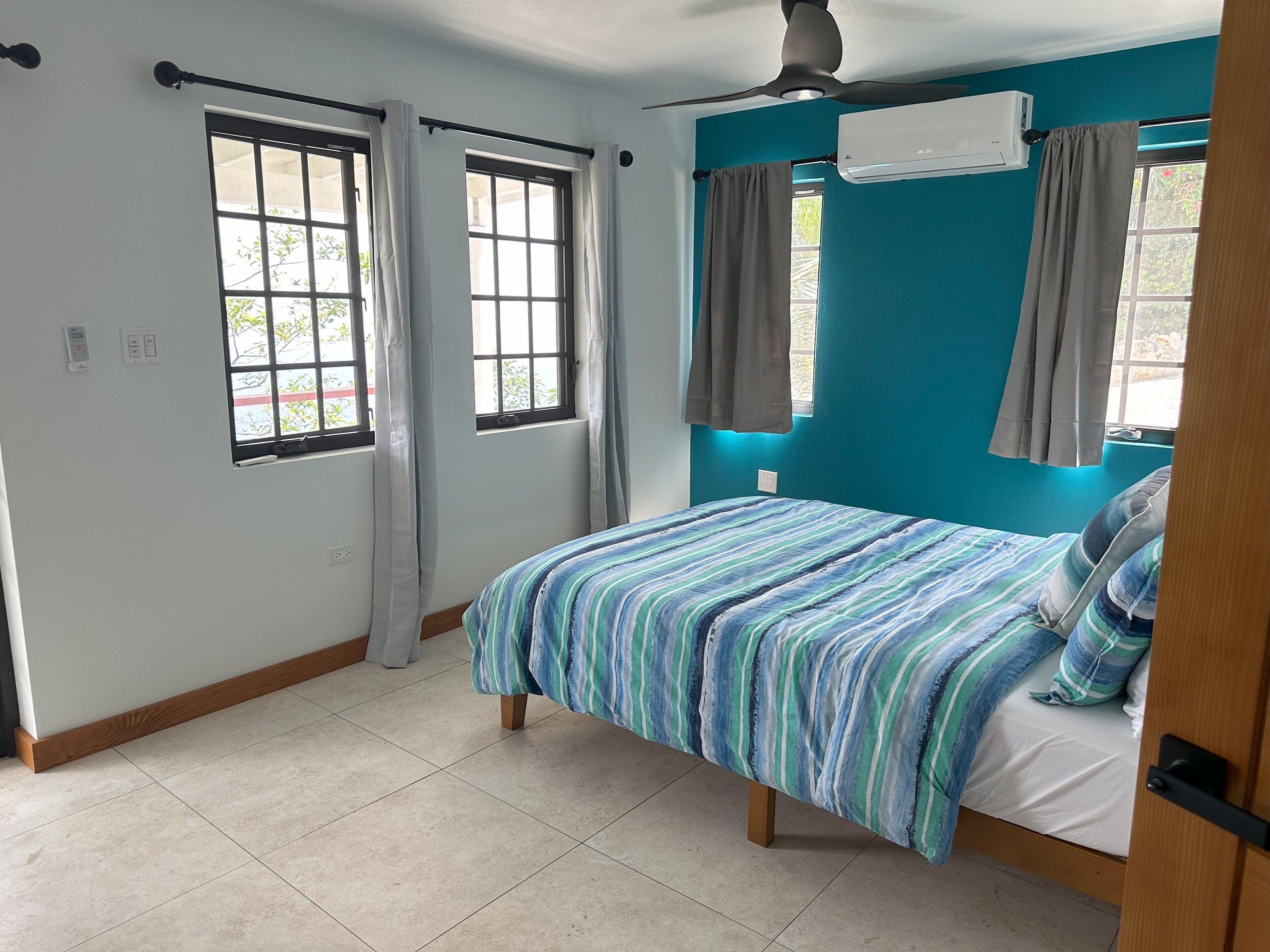 A bedroom with a bed, two windows with curtains, an air conditioning unit, and a ceiling fan.