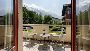 Un balcon avec deux chaises et une table, une bouteille de vin et un verre. On aperçoit, au loin, une montagne dont les sommets sont couverts de neige.