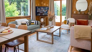 Un salon chaleureux avec un plancher en bois, une table à manger recouverte d’une nappe blanche, un canapé et un fauteuil. Il y a une grande fenêtre avec des rideaux, une bibliothèque et un miroir au mur.