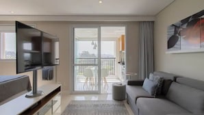 A modern living room with a flat-screen TV, grey sofa, and a balcony with outdoor seating. - Beautiful Studio - GRU 1112 (Jardim Flor da Montanha Guarulhos)