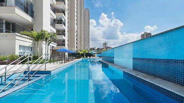 A swimming pool with a blue tiled wall, surrounded by a white building with balconies. - Beautiful Studio - GRU 1112 (Jardim Flor da Montanha Guarulhos)