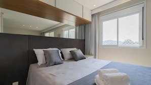 A modern hotel room with a large bed, a headboard, a window with curtains, and a view of the outside. - Beautiful Studio - GRU 1112 (Jardim Flor da Montanha Guarulhos)