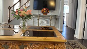 Een woonkamer met een houten salontafel, een tafel met een glazen blad, een aan de muur bevestigde televisie, een trap en een deur naar een andere kamer.