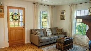Una sala de estar con piso de madera, un sofá gris con cojines, un baúl de madera y una ventana con cortinas.