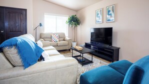 Ein Wohnzimmer mit einem beigen Sofa, einem blauen Sofa, einem Fernseher auf einem schwarzen Standfuß, einer Pflanze und zwei gerahmten Bildern an der Wand.