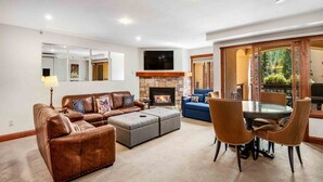 A living room with a fireplace, leather sofas, a dining table with chairs, and a large window with a view of greenery. - Ski In, Ski Out 1 Bedroom at the Eagle Bahn Gondola in Lionshead (Vail)