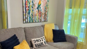 Ein Wohnzimmer mit einem grauen Sofa, gelben und blauen Kissen, einem farbenfrohen abstrakten Gemälde und einem Teppich mit Blumenmuster.