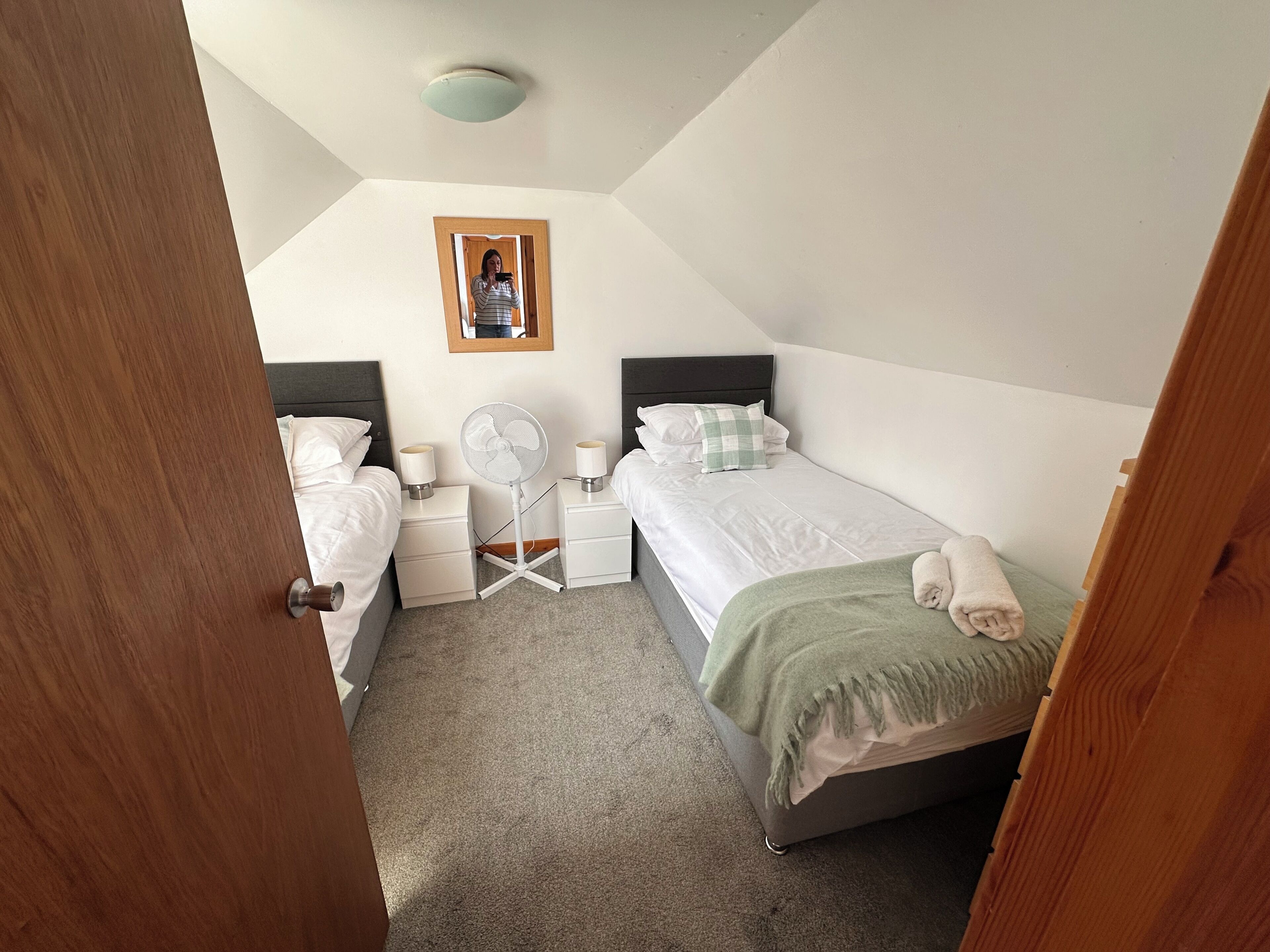 Quarto com duas camas, mesas de cabeceira, um ventilador e um quadro na parede, além de uma porta que dá para outro cômodo.