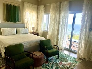 A bedroom with a large bed, two green armchairs, a small ottoman, a nightstand with a lamp, and a view of the ocean through sheer curtains.