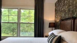 A hotel room with a large window, a bed with patterned bedding, a nightstand with a lamp, and a decorative wall panel.