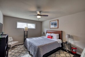 A bedroom with a bed, a desk, a chair, a standing lamp, a ceiling fan, a window, and a framed picture on the wall.