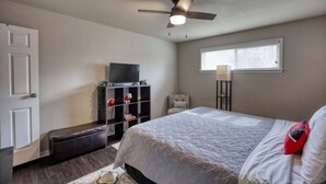 Une chambre à coucher avec un lit, un ventilateur de plafond, un téléviseur posé sur un meuble, une fenêtre et une porte.