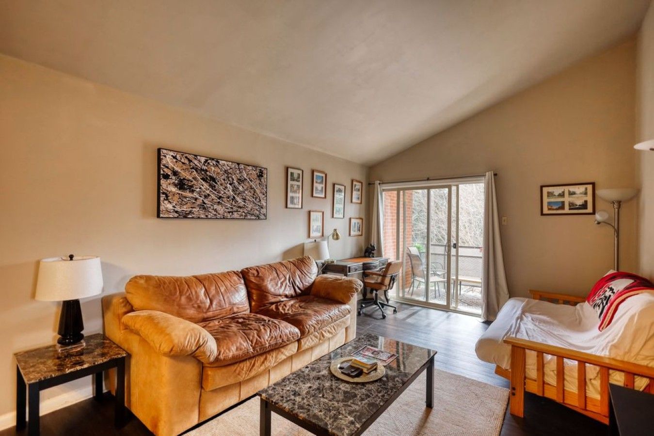 A living room with a brown leather sofa, a glass coffee table, a wooden bed, and a sliding glass door leading to a balcony.