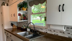 Una cocina con armarios blancos, un lavamanos de acero inoxidable, una ventana con vistas a un árbol y un césped, y un salpicadero de azulejos.