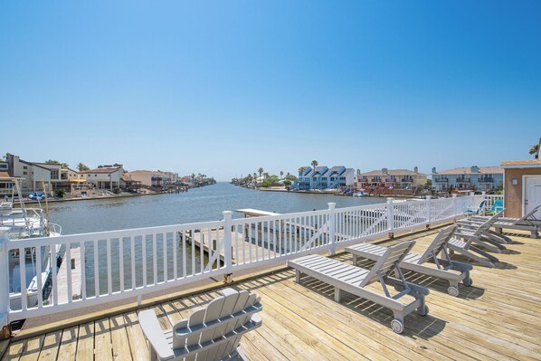 A wooden pier with white railings, lounge chairs, and a view of a marina and buildings across the water.