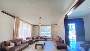 A spacious living room with a large sectional sofa, a wooden coffee table, and a view of the outdoors through large windows. - Jim Holiday Homes (Mombasa)