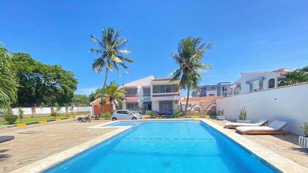A swimming pool with lounge chairs, palm trees, and a building with a tiled roof in the background. - Jim Holiday Homes (Mombasa)
